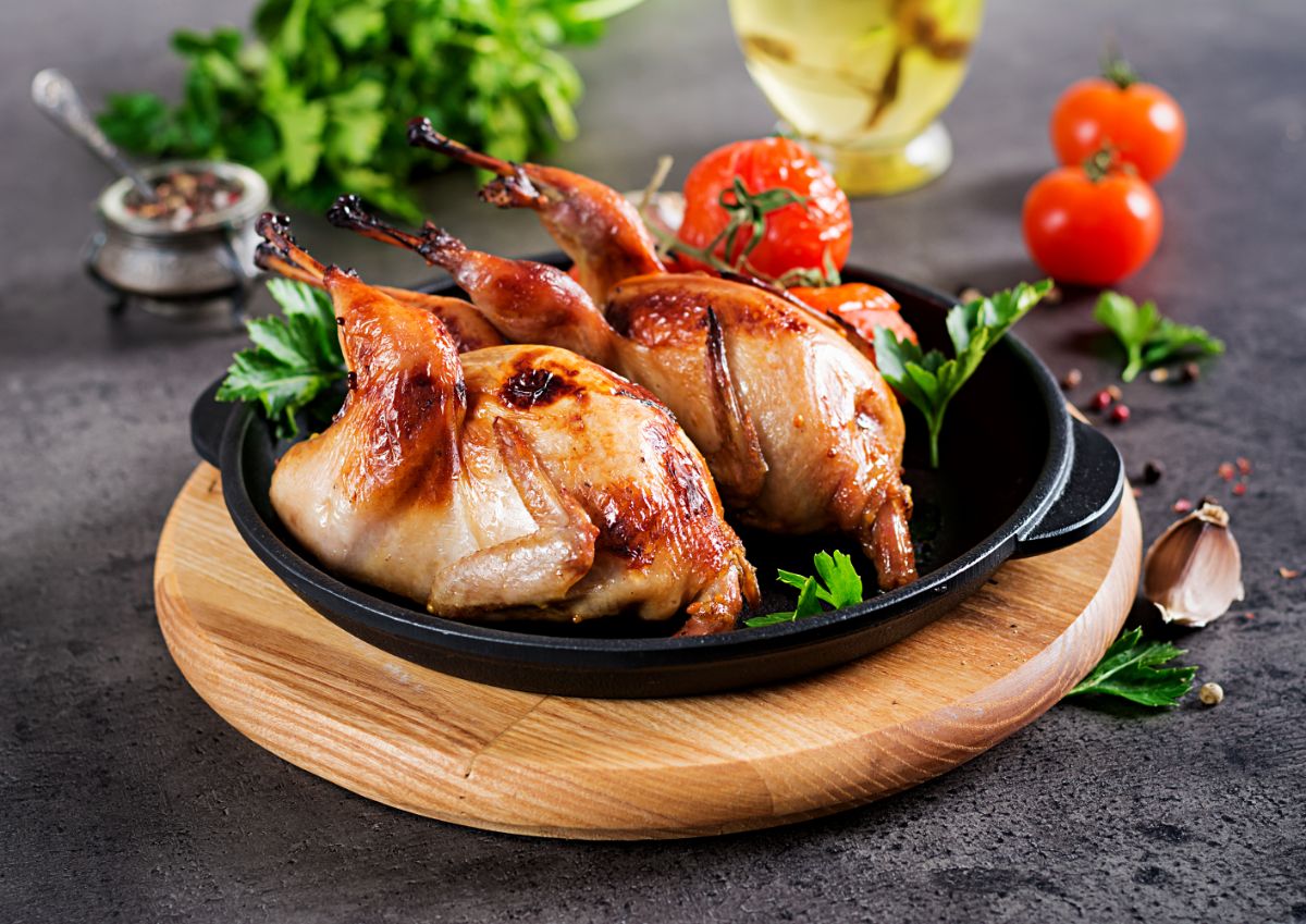 Two baked quails on a black pan on a wooden board on a table with vegetables.
