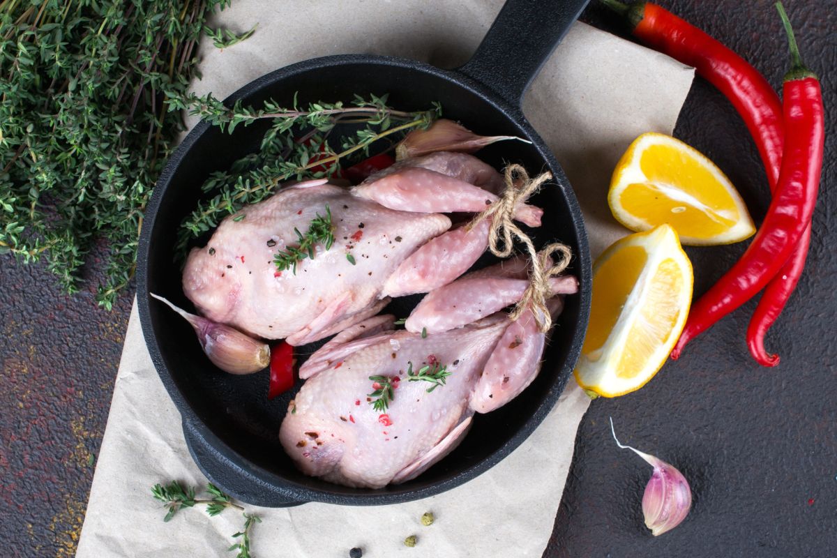 Two raw quails in a black pan with vegetables, herbs and sliced lemons on a table.