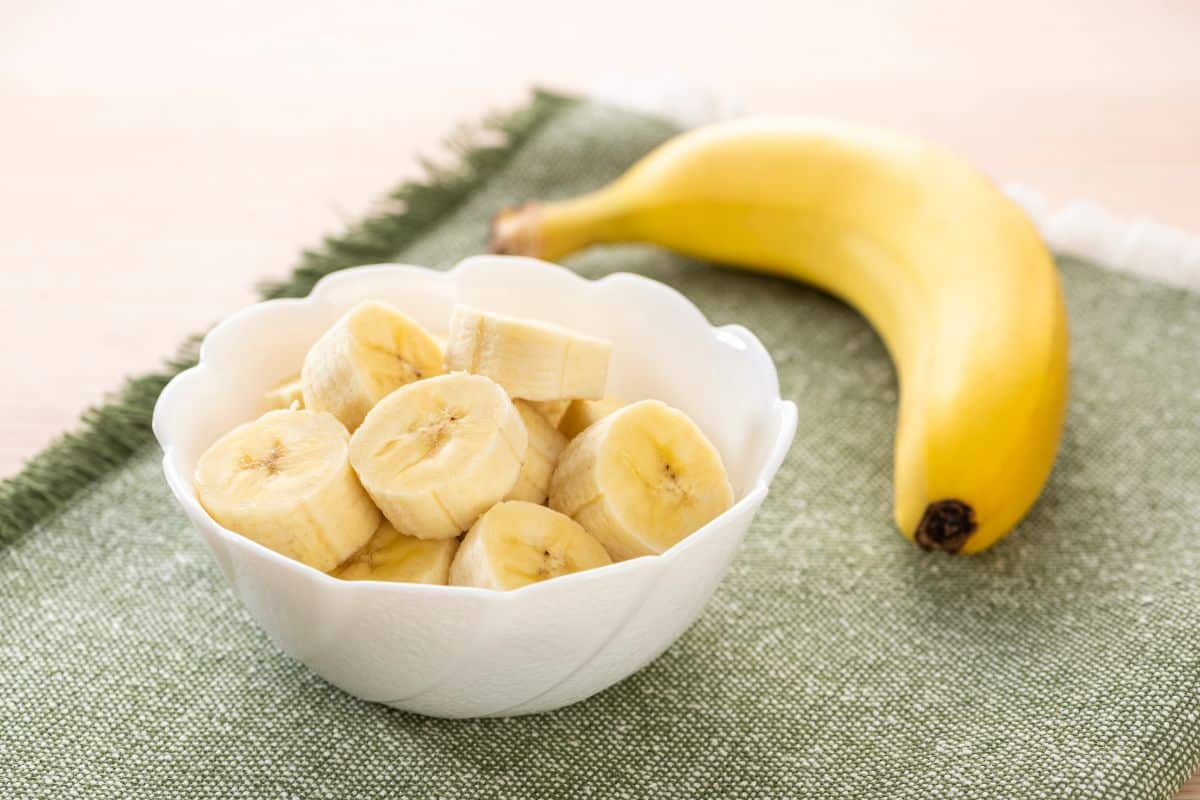 A white bowl with sliced bananas on a table next to a whole banana.,