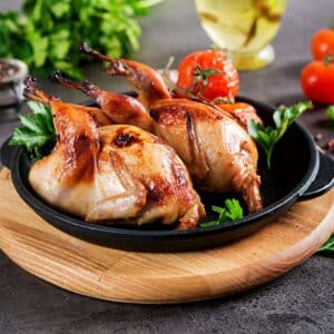 Two baked quails on a black pan on a wooden board.
